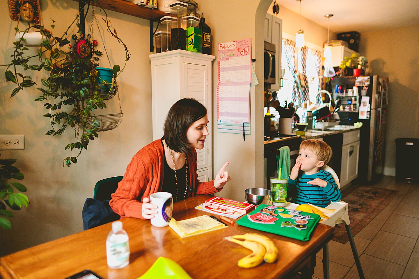 Kid eating a banana at kitchen table while talking to his mom