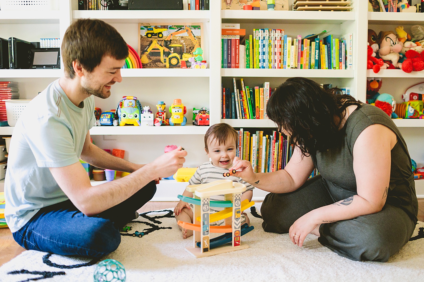 Baby sitting in front of library bookcase playing with toy cars with his mom and dad