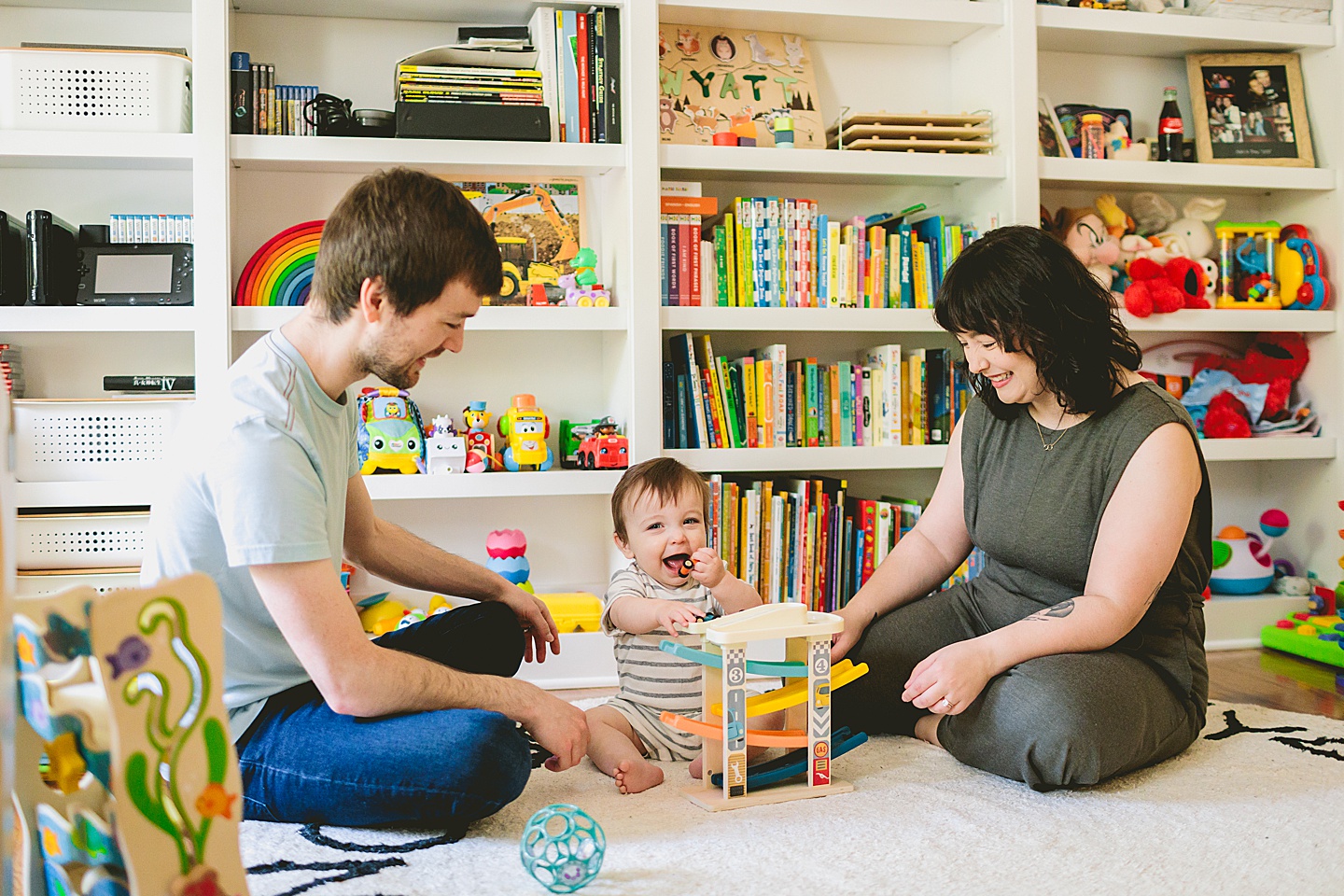 Baby sitting in front of library bookcase playing with toy cars with his mom and dad