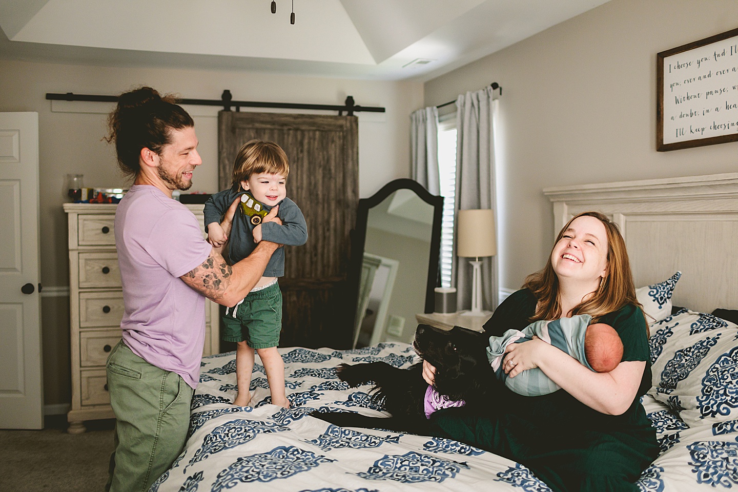 Toddler jumping on the bed with dad while Mom sits with baby and dog