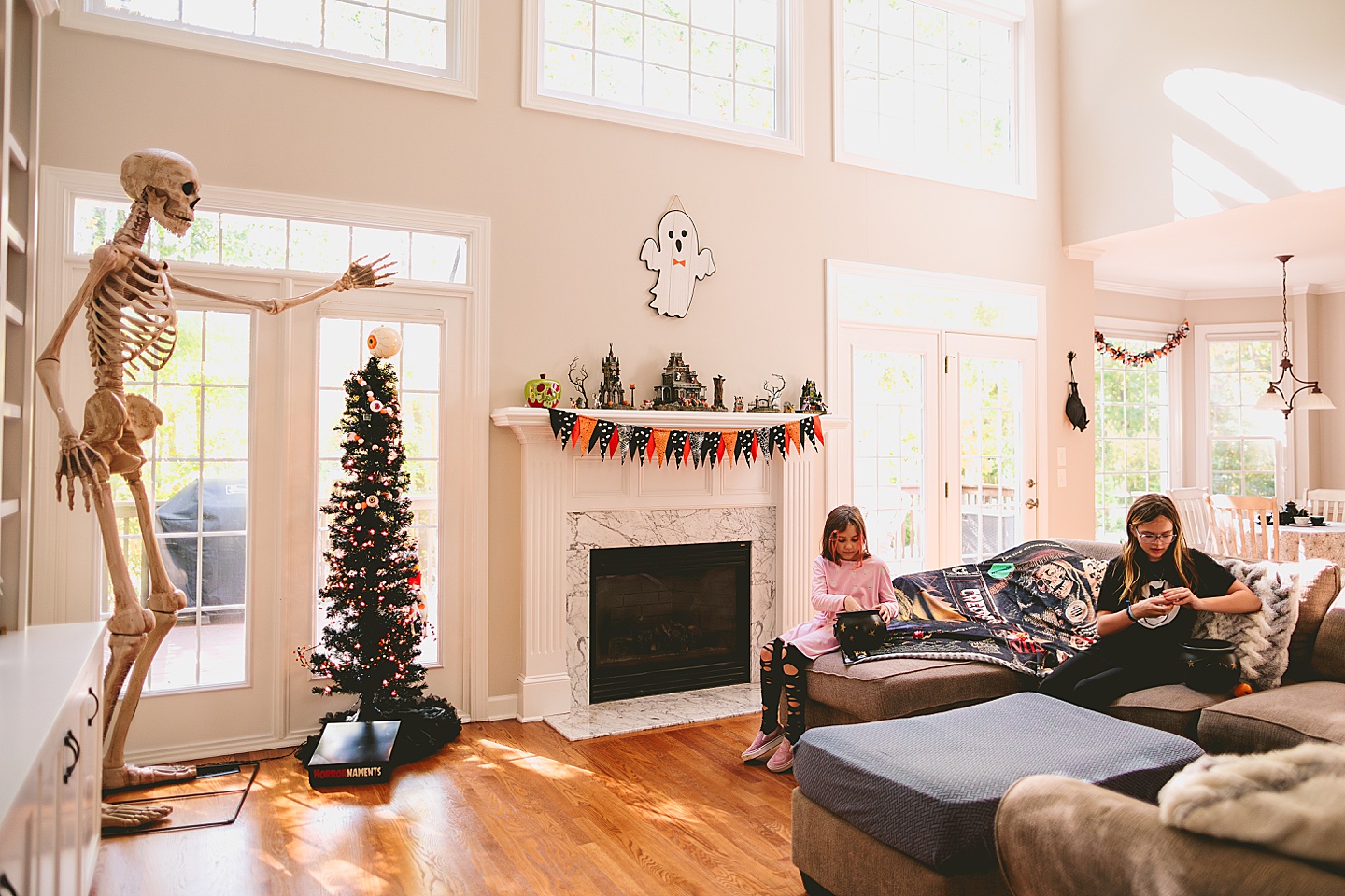 Sisters sit on a couch next to a fireplace and a giant skeleton and open Halloween eggs