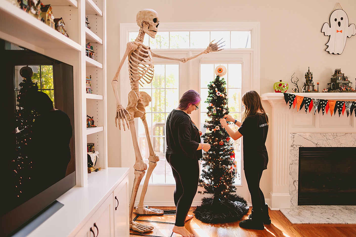 Mother and daughter decorate black Halloween tree with horror ornaments next to eight foot tall skeleton in living room