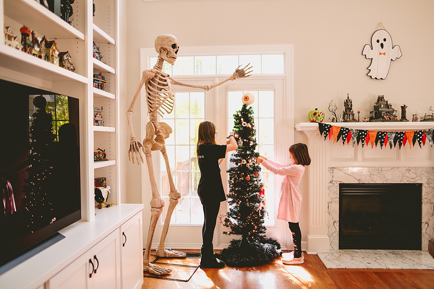Sisters decorate black Halloween tree with horror ornaments next to eight foot tall skeleton in living room