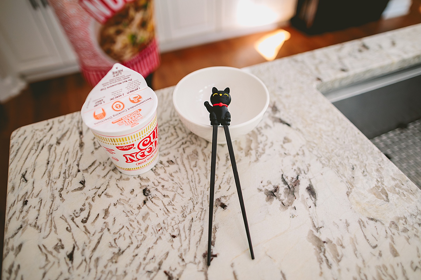 Cup of Noodles sits next to a bowl with cat chopsticks leaning on it