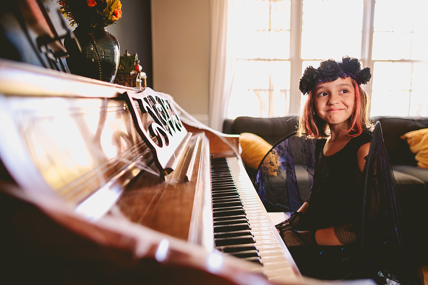 Girl in a dark fairy costume with black wings sits next to the piano and smiles while wearing black flower crown