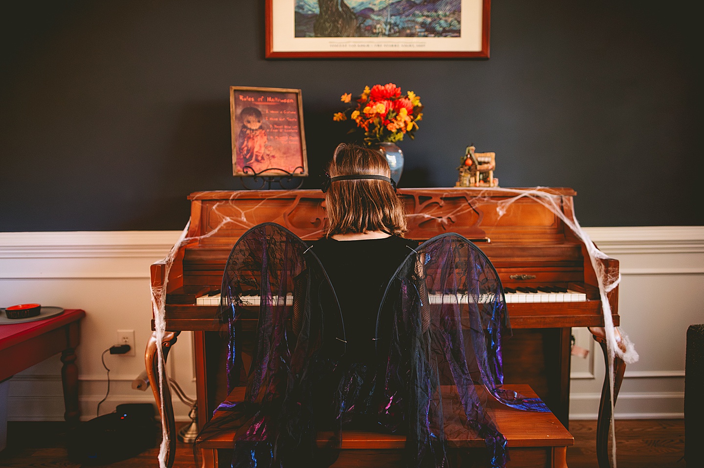 The back of a girl wearing dark fairy wings playing piano in a living room that's decorated for Halloween