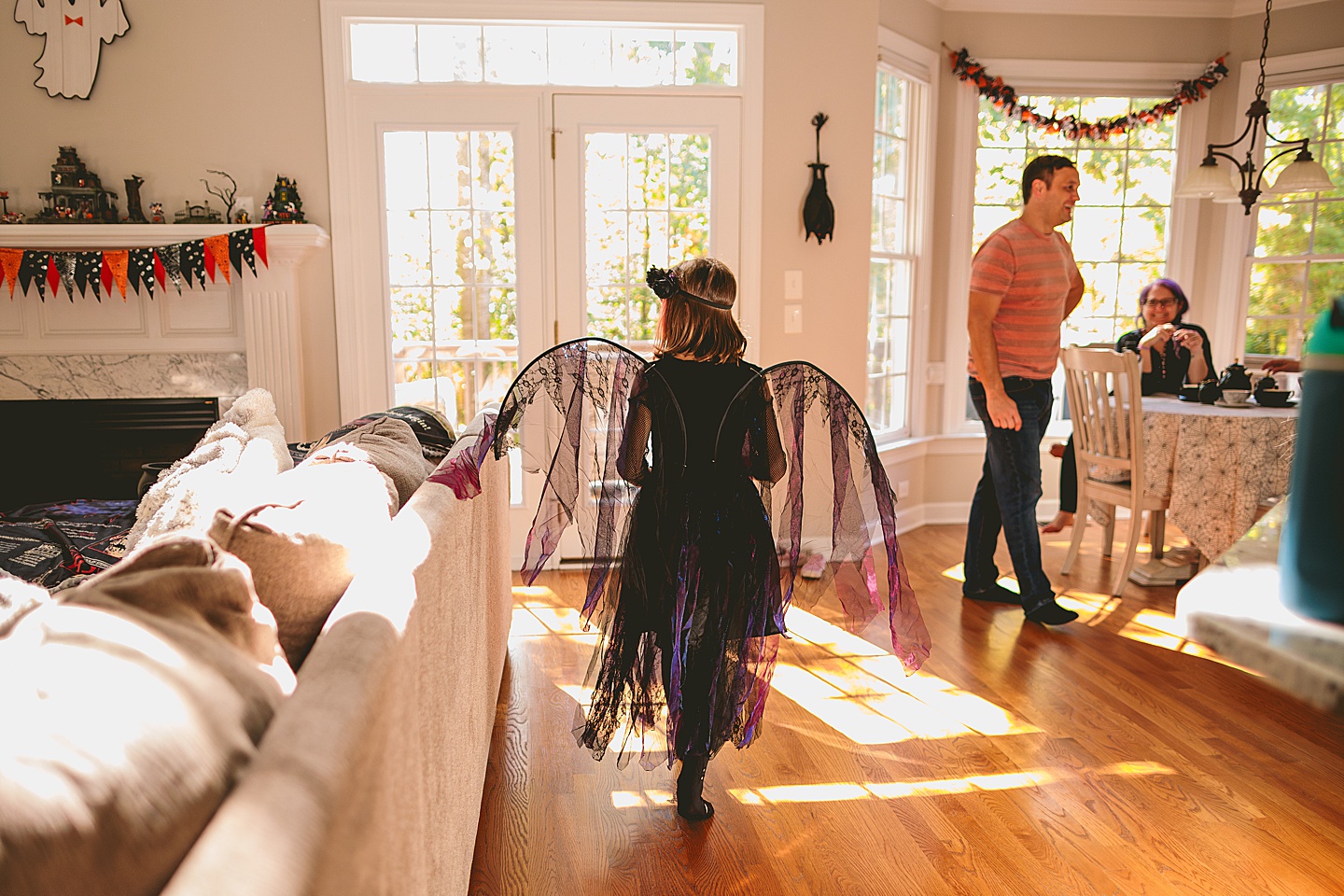 Girl wearing a dark fairy costume with black wings walks through a sunlit living room